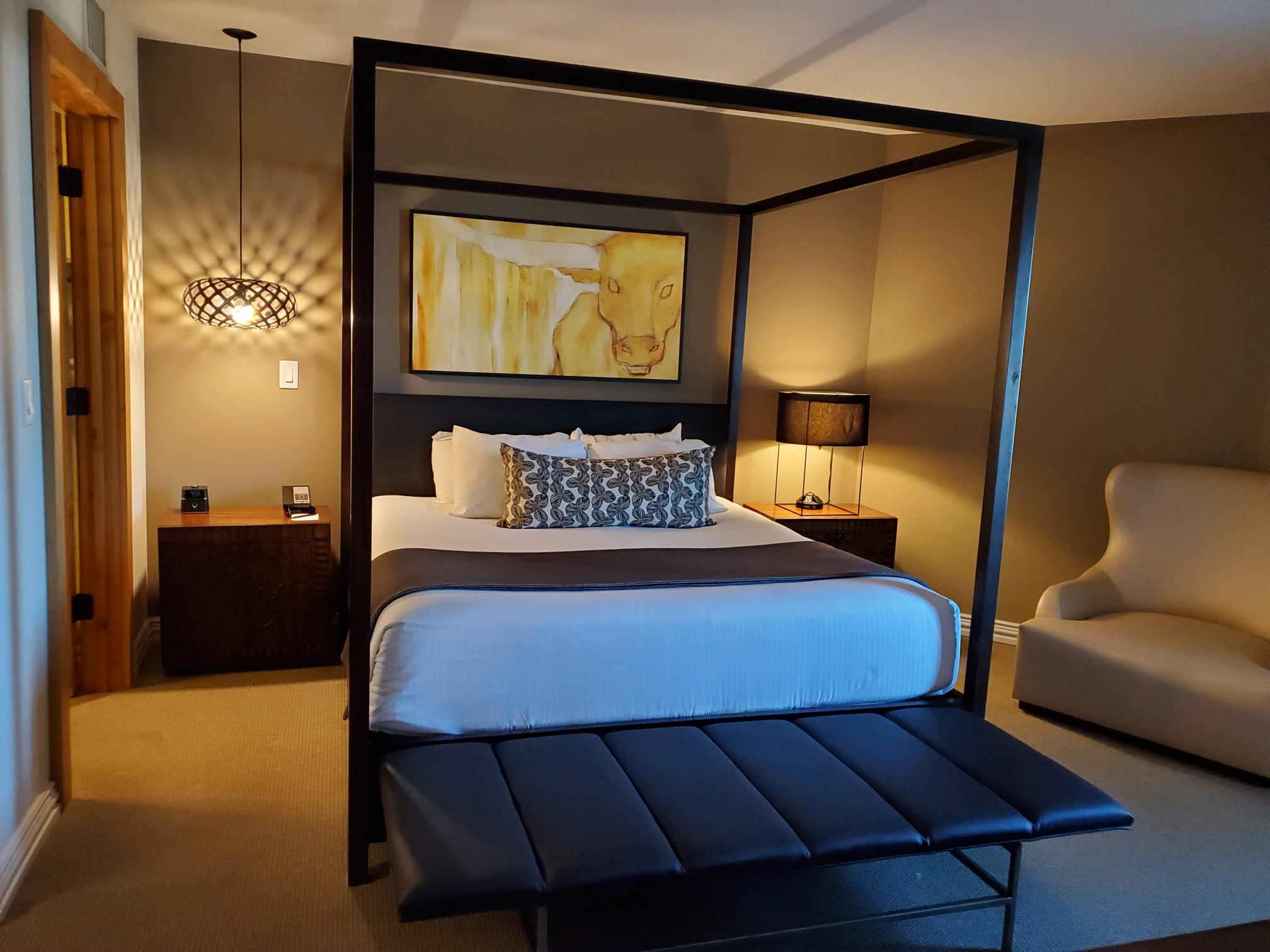 A wide view of a hotel room with a four-poster bed, bench, nightstands, and artwork.
