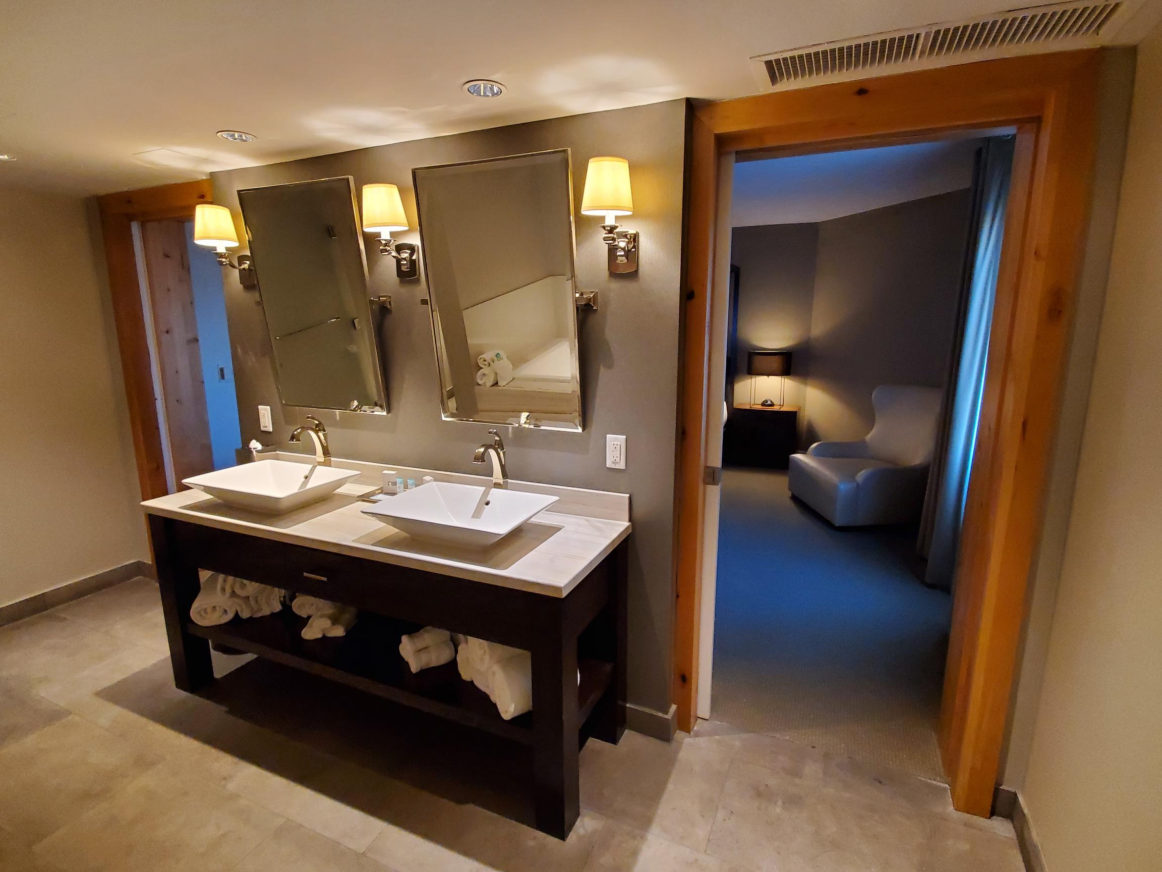 A modern bathroom with a double vanity, two sinks, and a doorway leading to a bedroom.