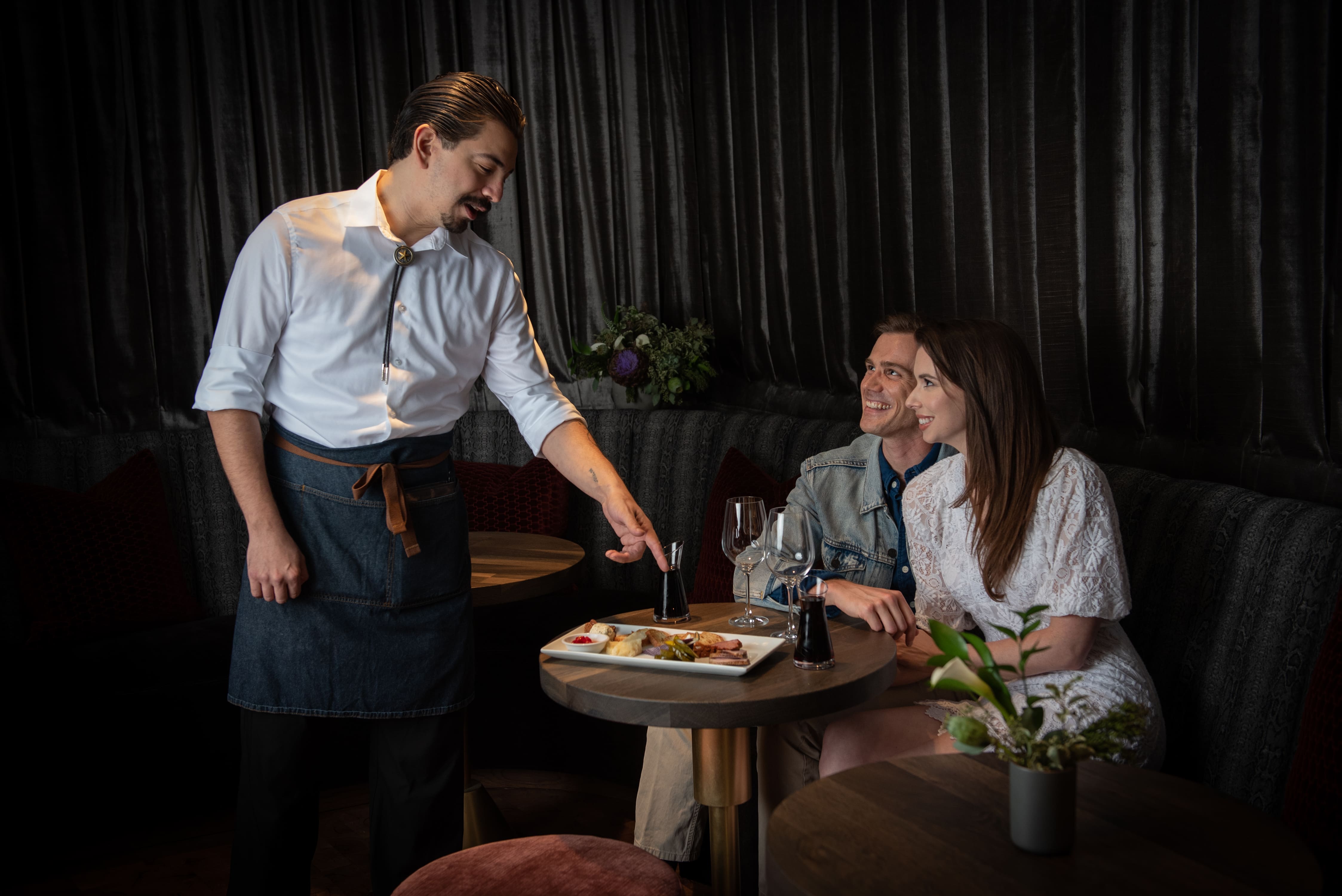 A waiter presenting a dish to a couple sitting at a table in a dimly lit restaurant.