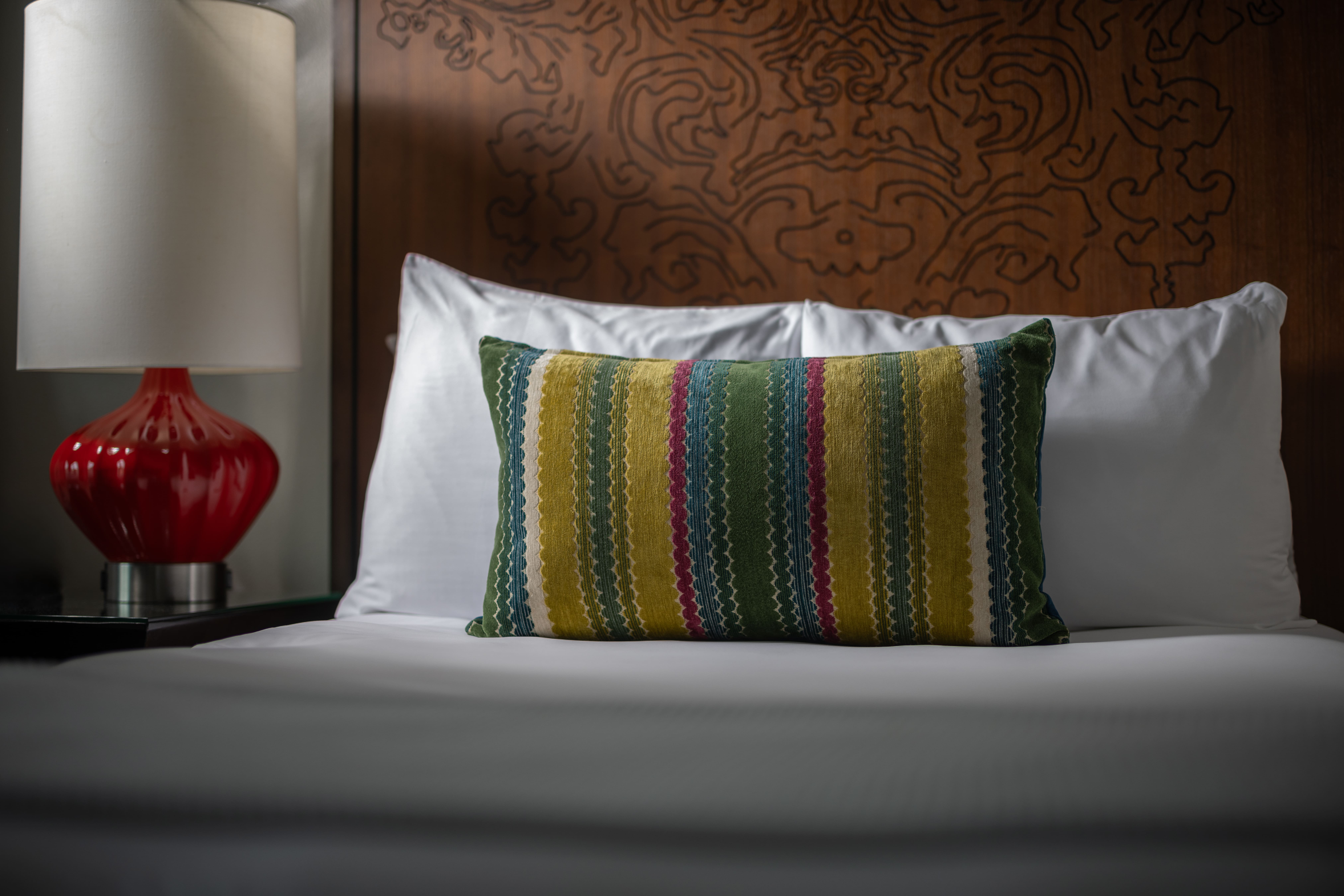 A close-up of a bed with white bedding, a colorful pillow, and a red lamp on a nightstand.