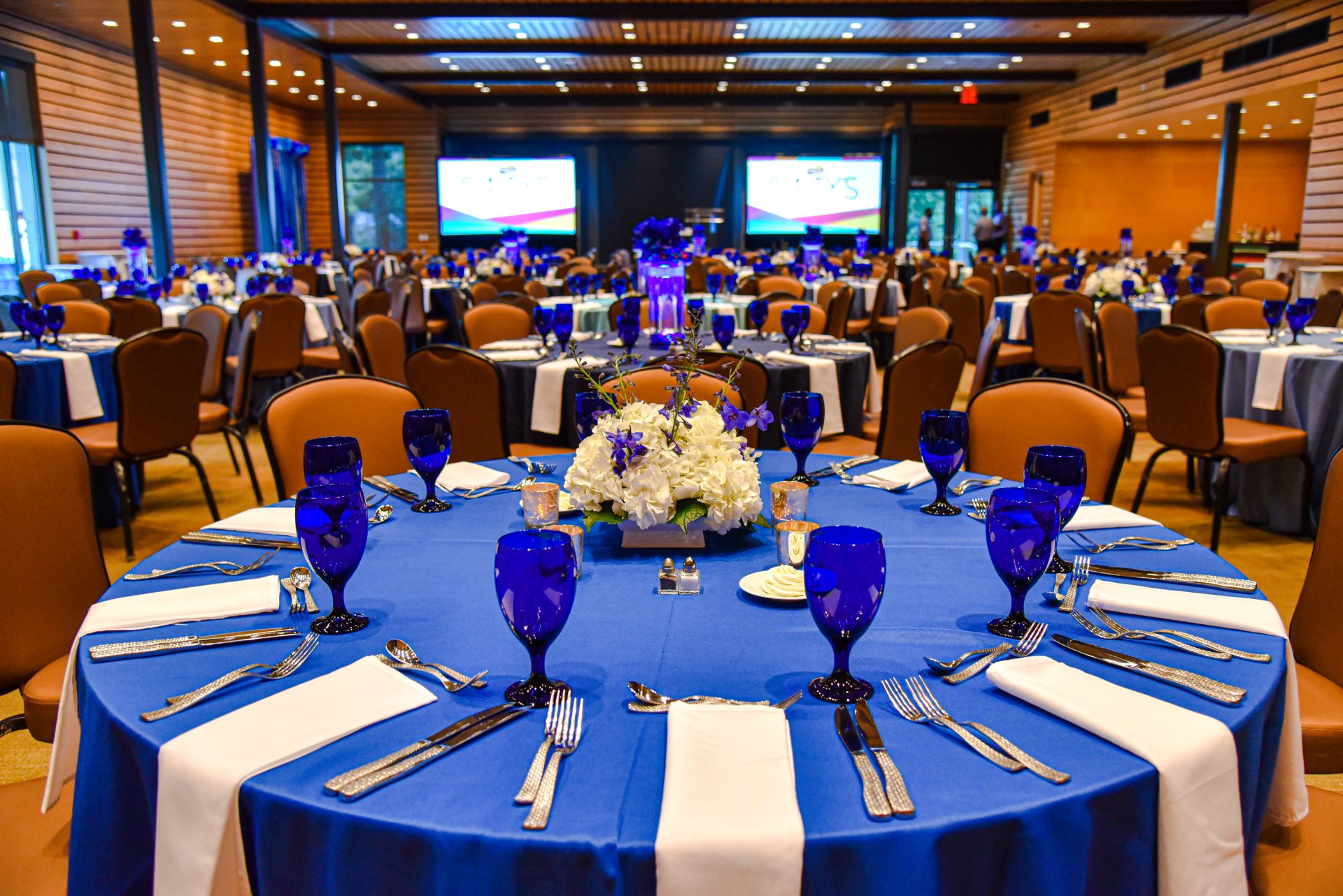 A large, formally set dining room with round tables, blue tablecloths, and floral centerpieces.