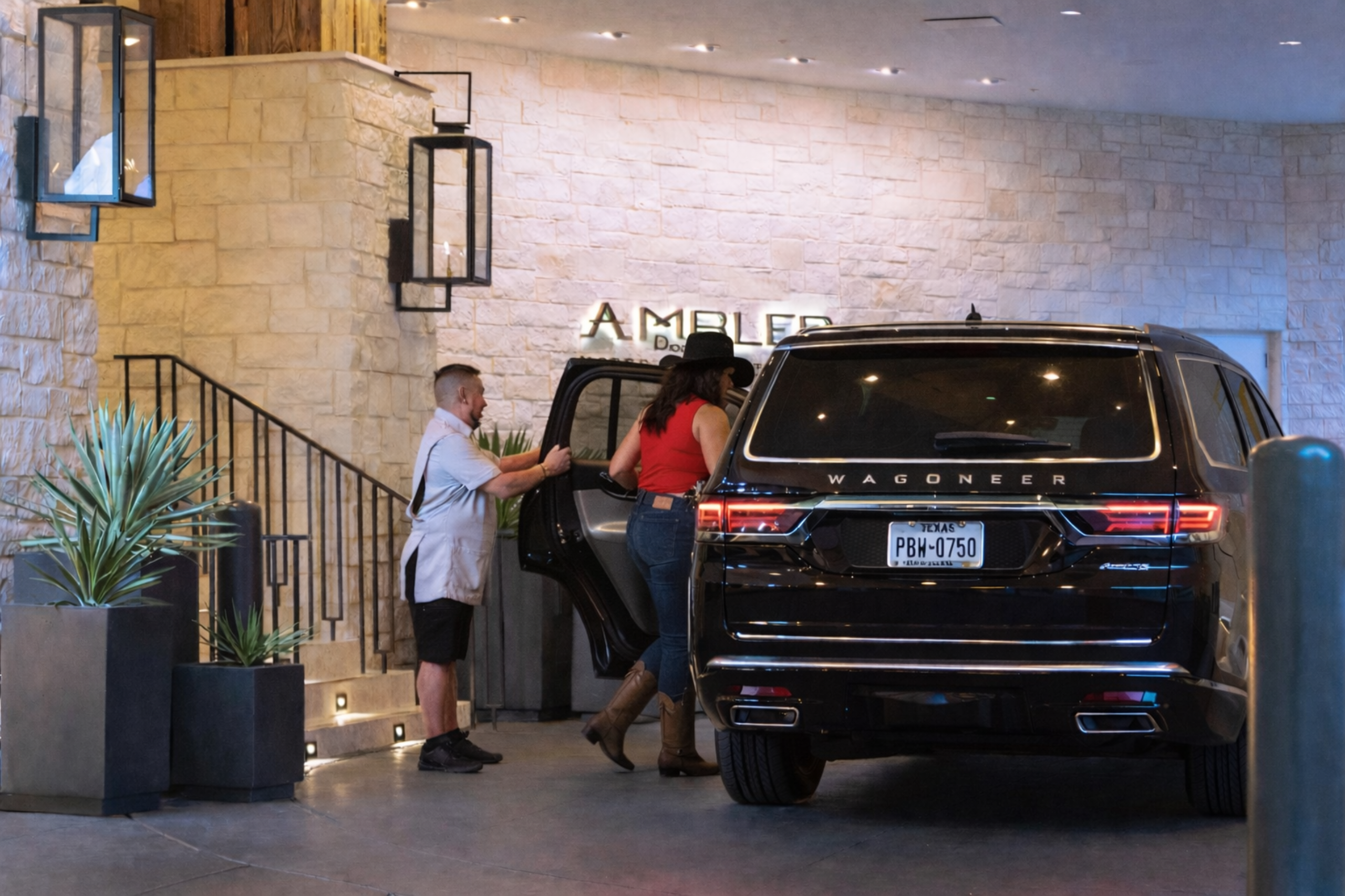 A man is helping a woman out of a black SUV in front of a stone wall with a staircase.