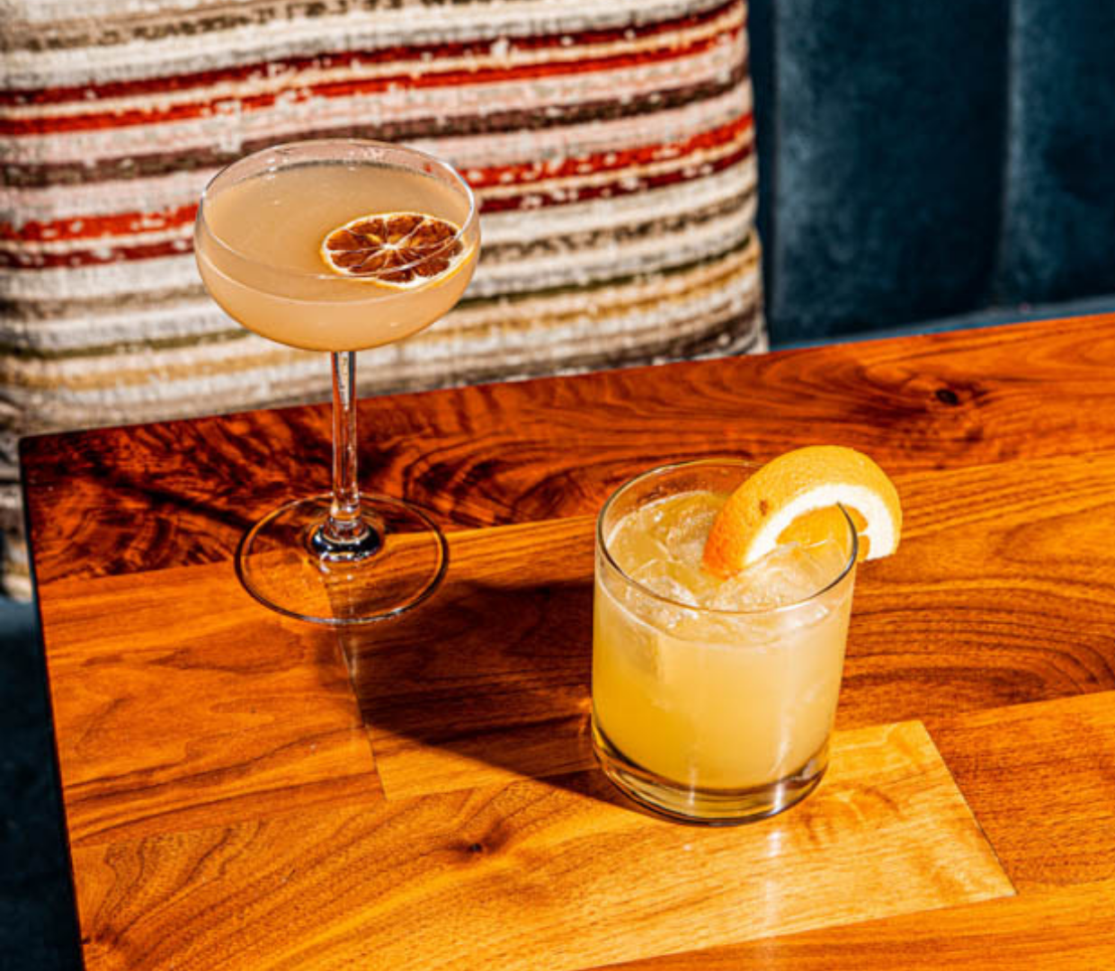 Two cocktails on a wooden table with a striped cushion and blue velvet chair